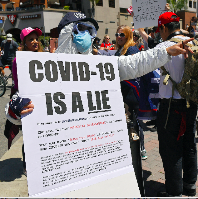 Person wearing a mask and holding a sign with the heading, &quot;COVID-19 is a lie,&quot; followed by text addressing statements by CNN and the CDC