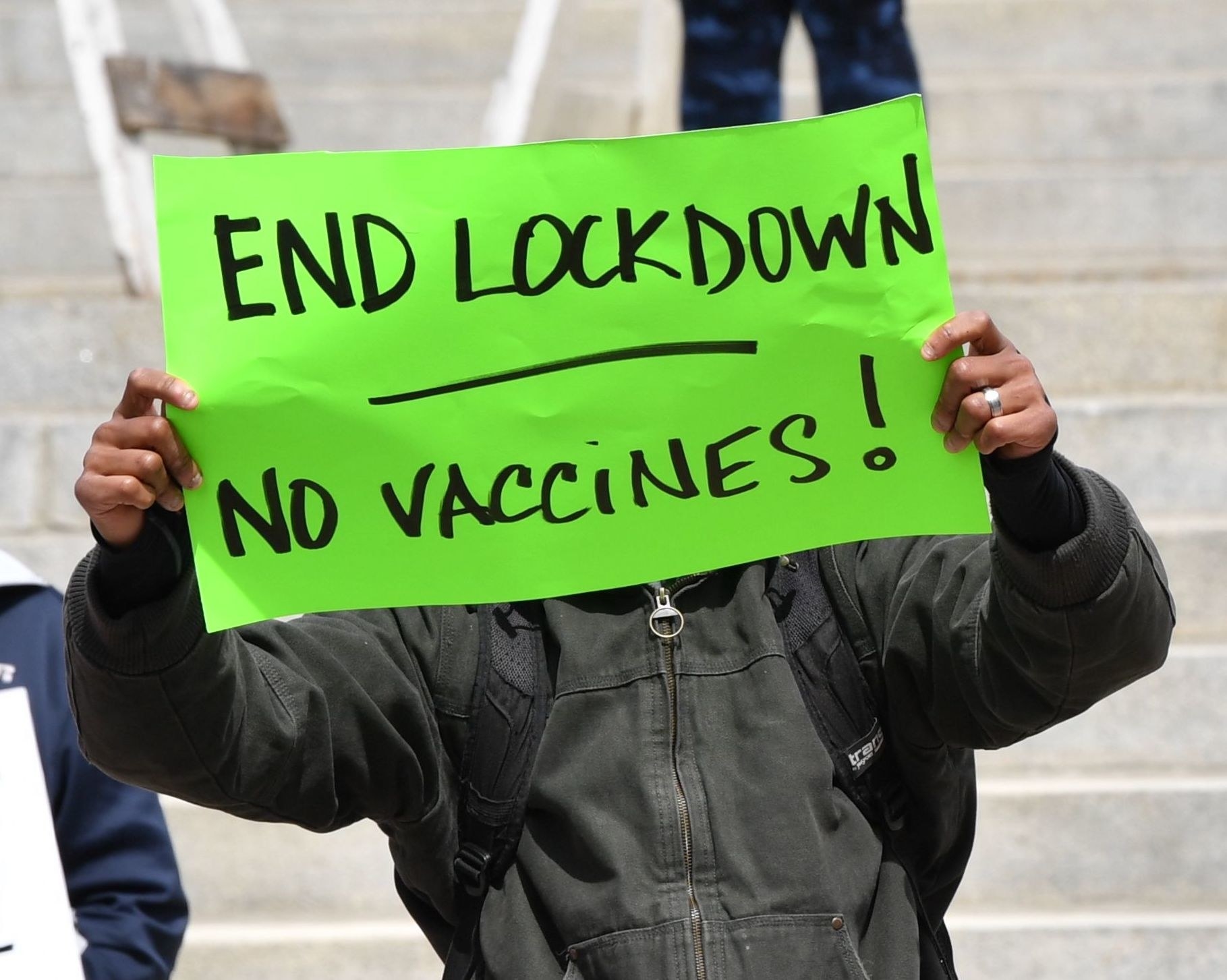 Person holding a sign in front of their face reading, &quot;End lockdown no vaccines!&quot;