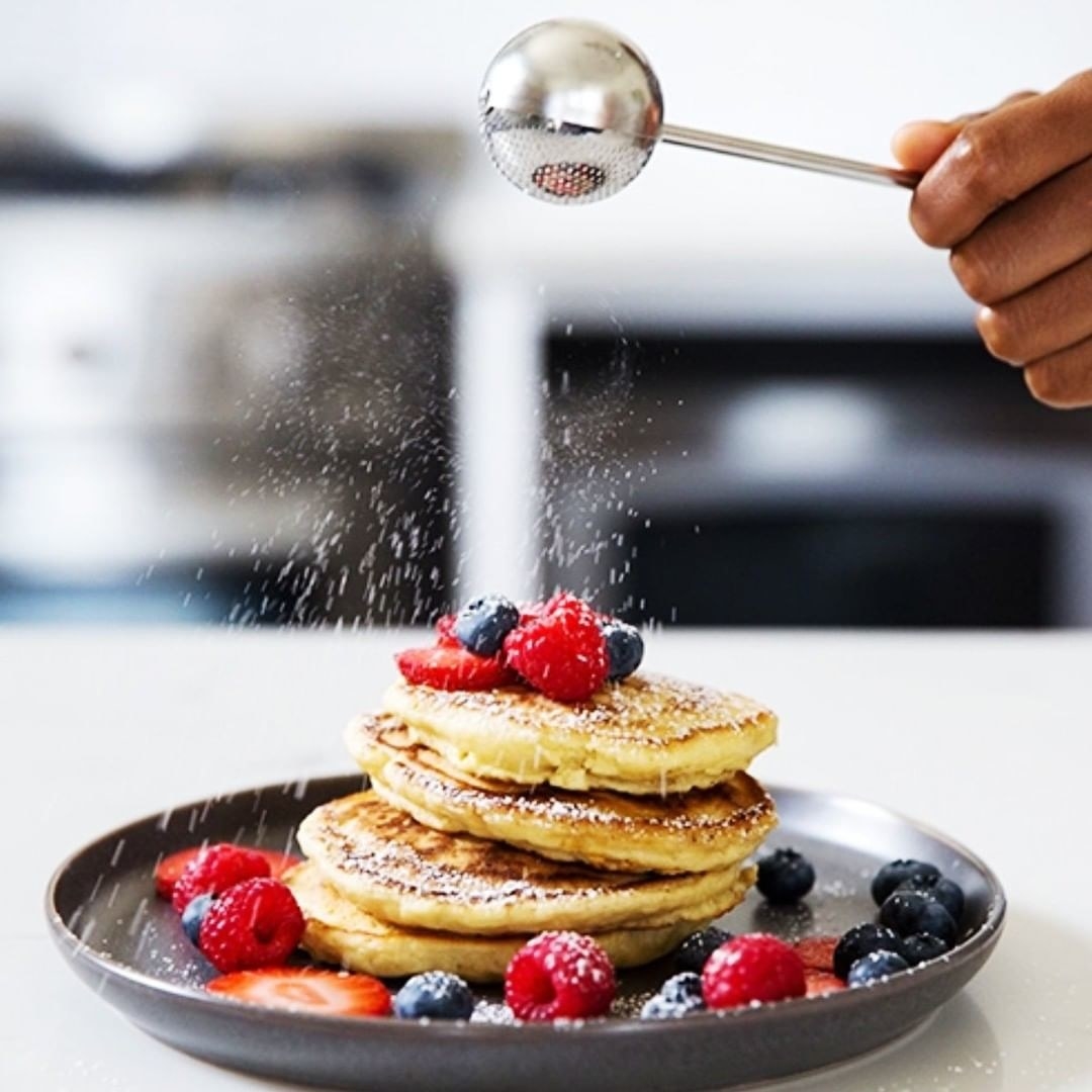 A wand is sprinkling powdered sugar over pancakes with berries