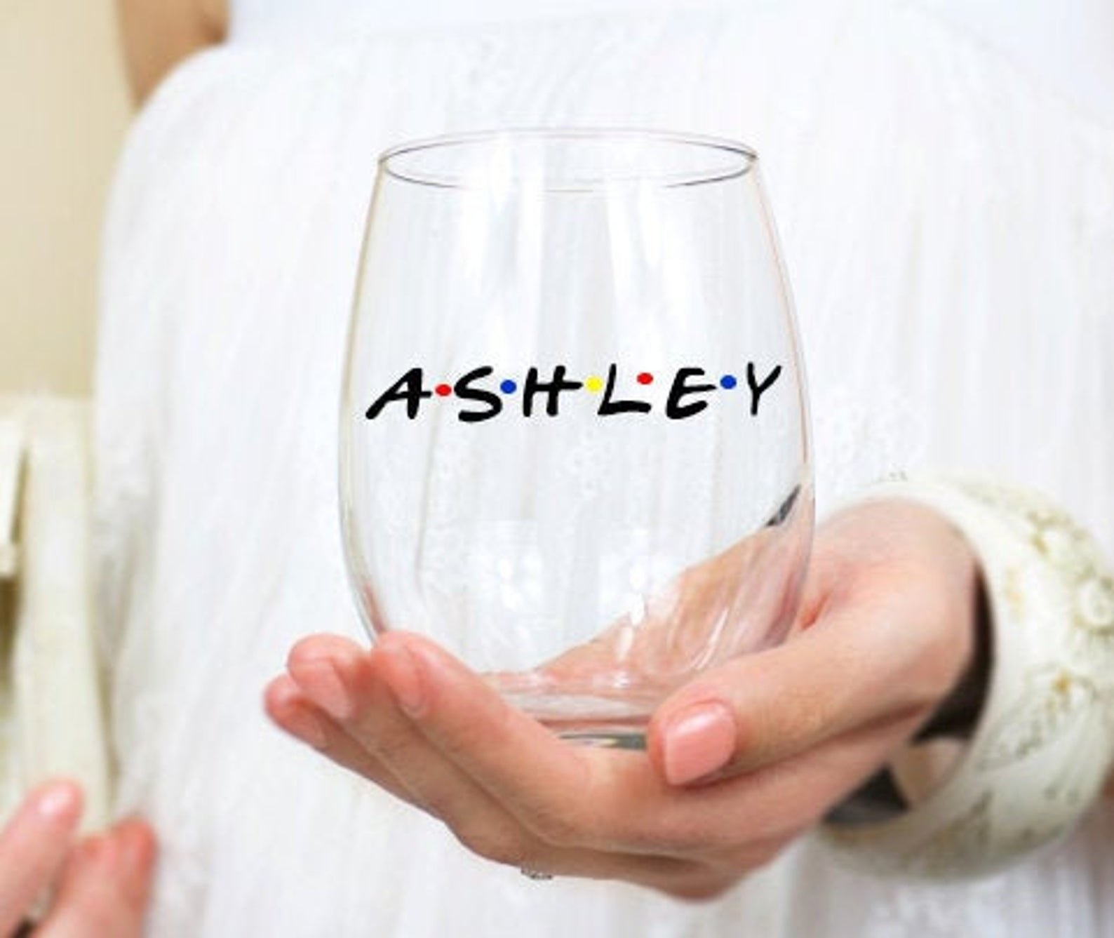 Person is holding a stemless wine glass with the name Ashley on it with dots in between each letter