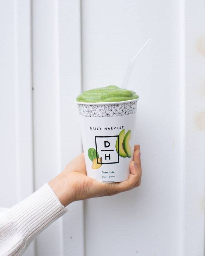 A hand holding a paper cup filled with a bright green smoothie from Daily Harvest. 
