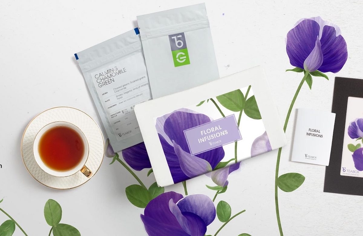 a single filled teacup in a saucer next to three packages of tea (one has pretty purple flower petals on it)