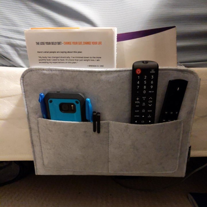 Reviewer showing the caddy attached to the side of a bed, holding remotes, a phone, and books