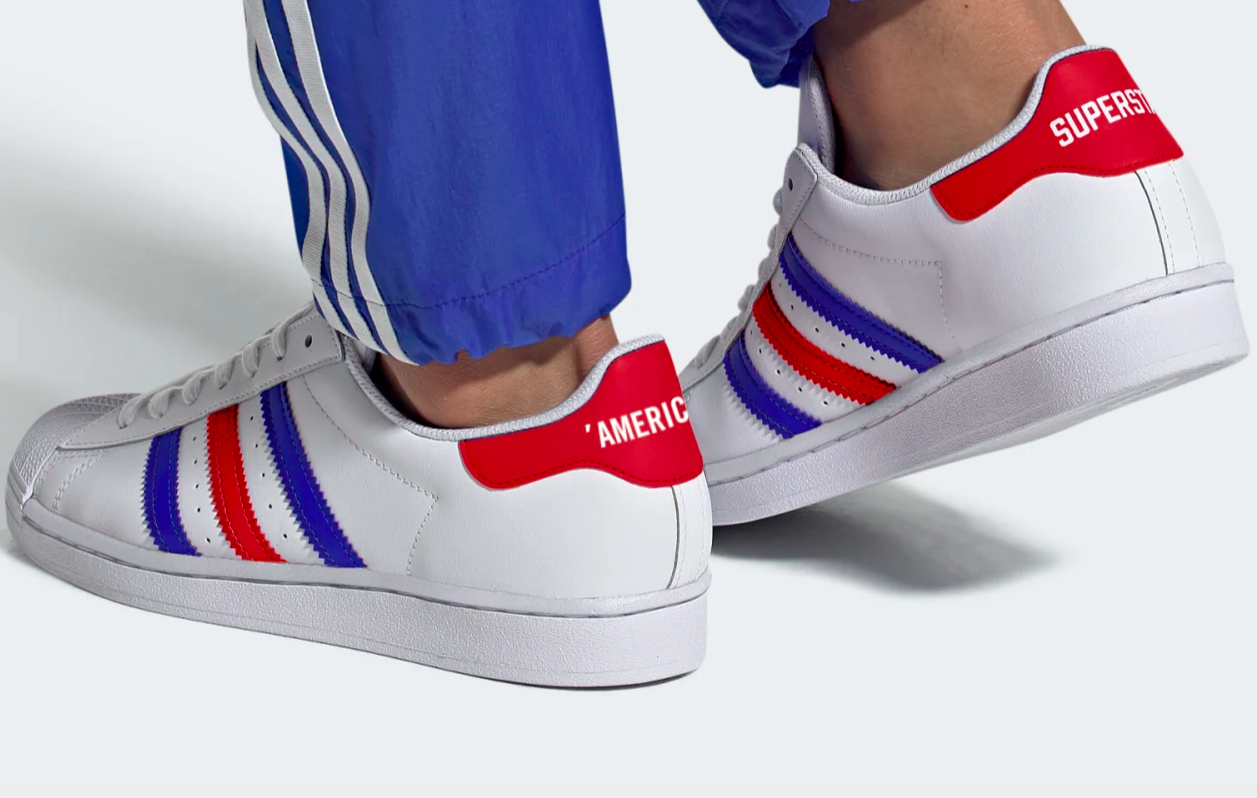 White shoe with blue and red alternating Adidas stripes down the center side with a bright red heal lip