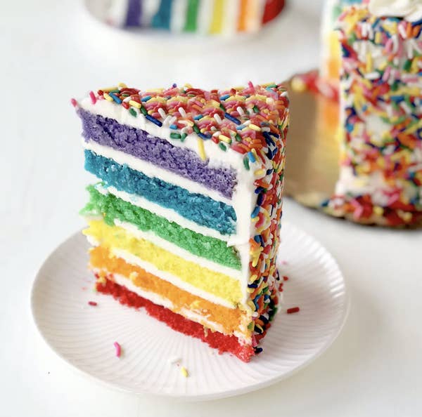 A slice of cake cut to show rainbow layers inside and rainbow sprinkles outside 