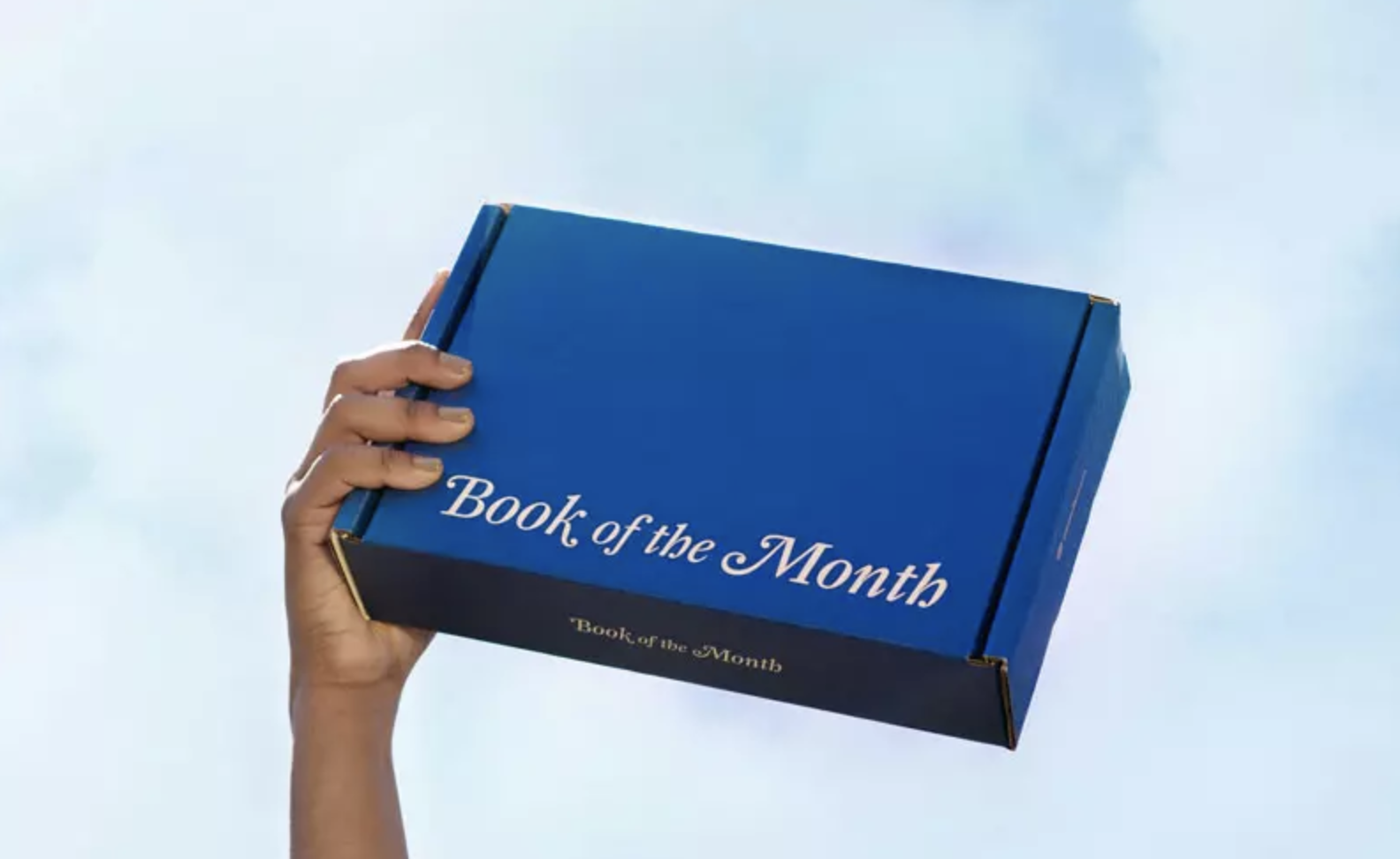 A cropped shot of a hand holding a blue Book of the Month cardboard subscription box in the sky