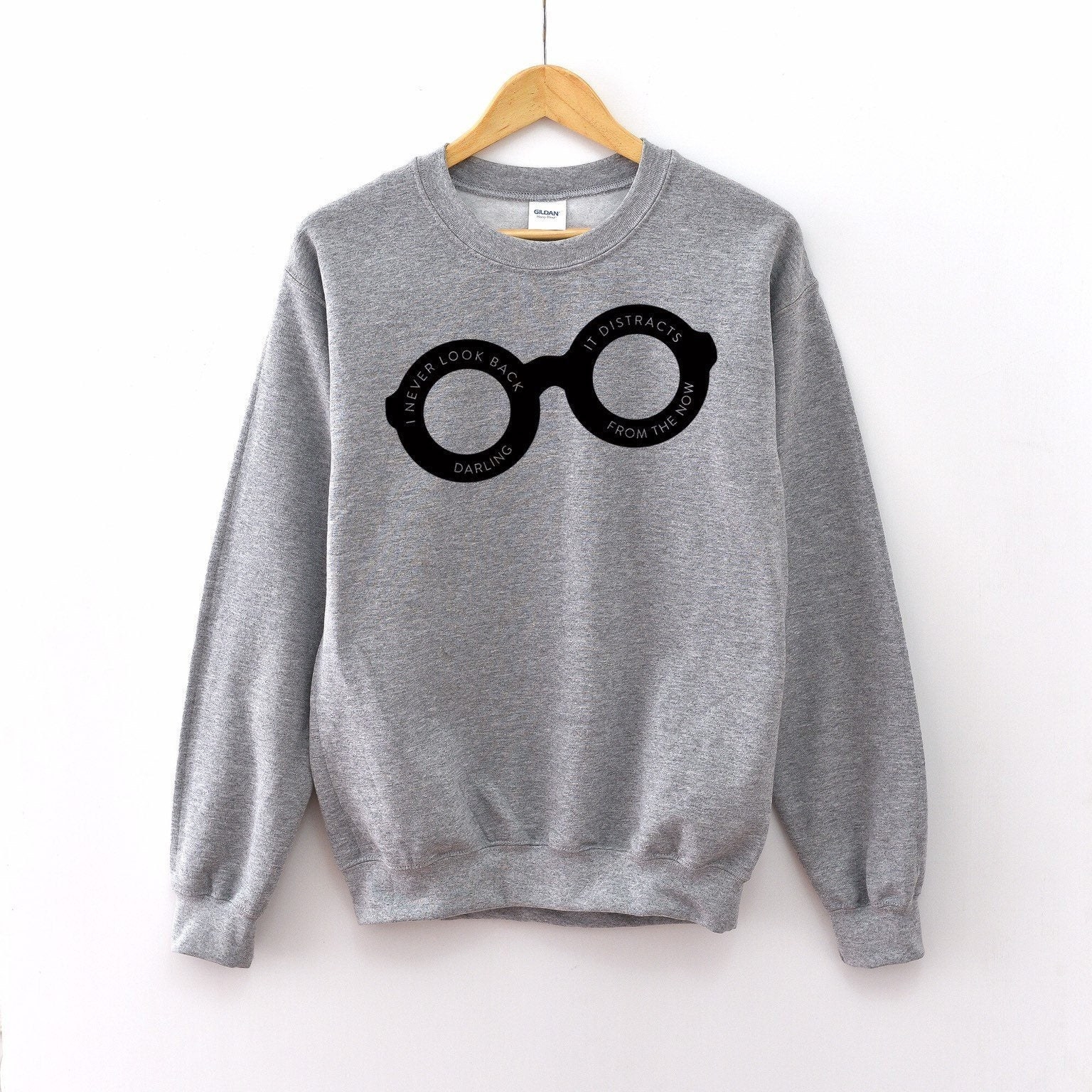 a grey pullover with black glasses on it that says &quot;i never look back darling it distracts from the now&quot; inside them