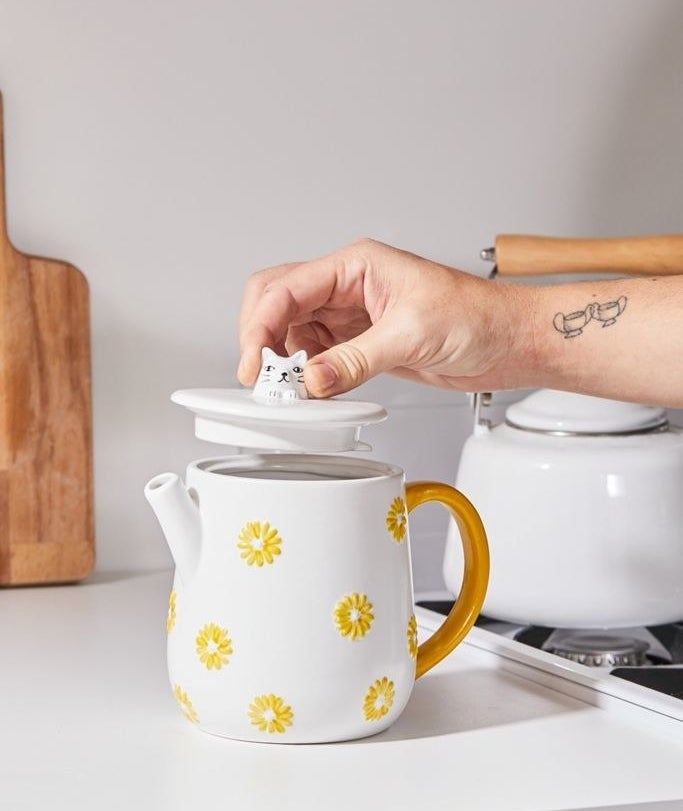 A hand lifting the lid of the white teapot with yellow flowers, a gold handle, and a cat head handle on the lid 