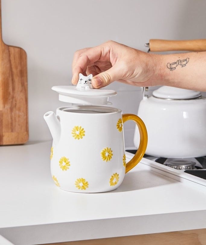A hand lifting the lid of the white teapot with yellow flowers, a gold handle, and a cat head handle on the lid 