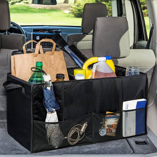 A black trunk organizer inside a car trunk filled with car accessories, groceries, an umbrella and more in its mesh side pockets