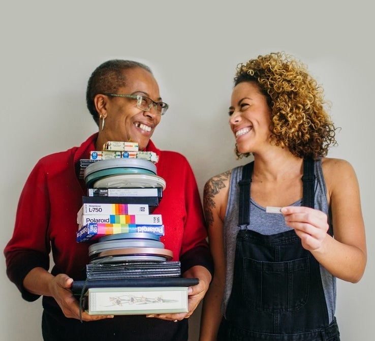 Two people standing next to each other: one older, holding a stack of photo albums, film reels, and homemade video cassettes, and one younger, holding a small USB drive