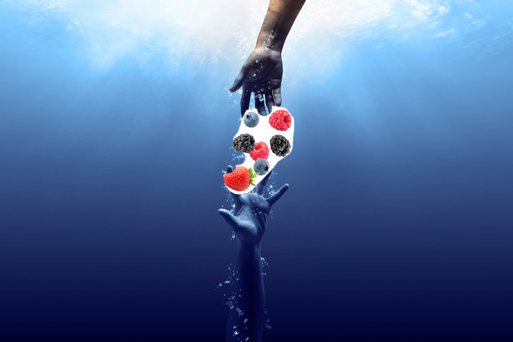 Hand dropping berries into water with splashes; berries appear suspended mid-fall above another hand underwater catching them