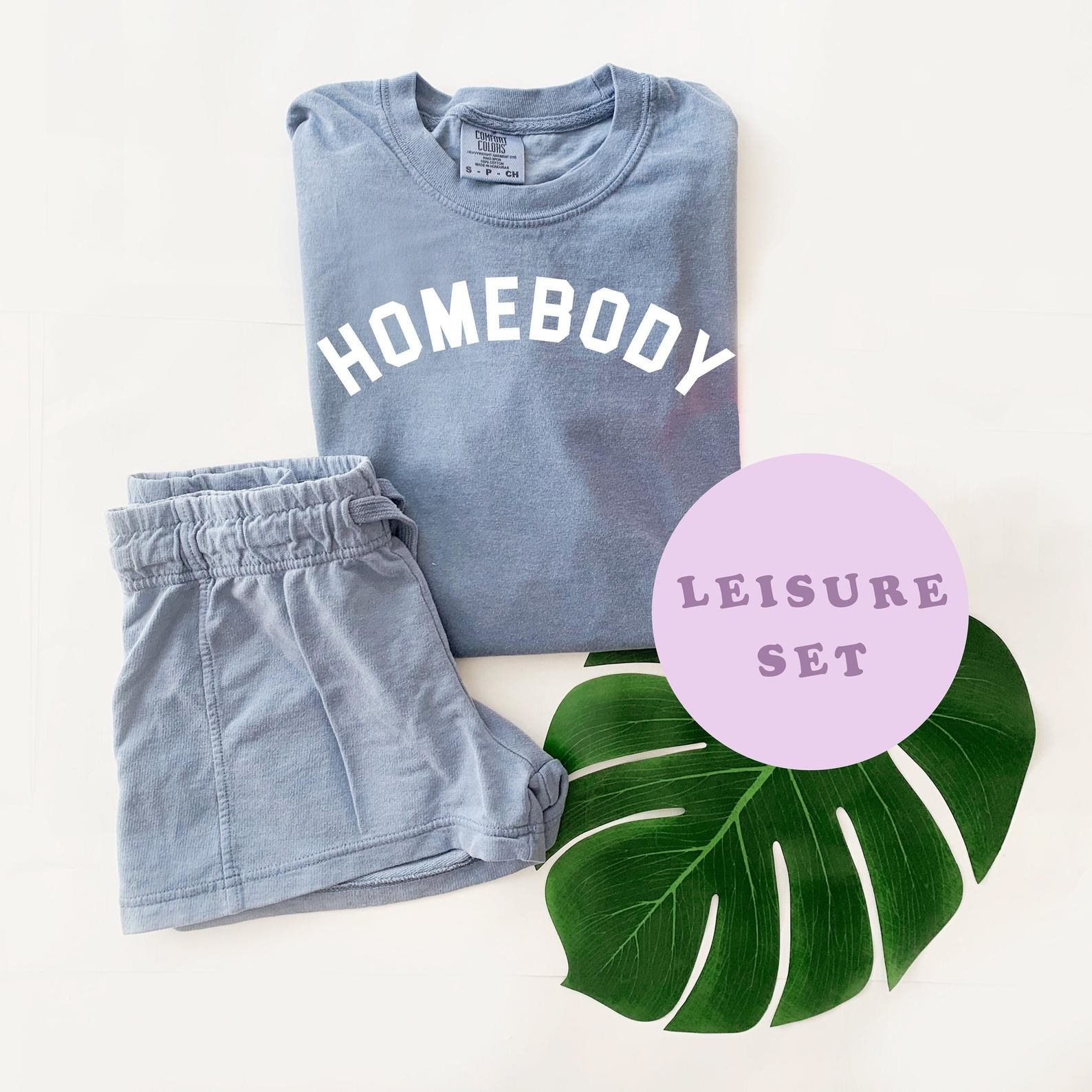 A folded pajama set with a pair of blue drawstring shorts and a matching blue long-sleeved shirt that says &quot;Homebody&quot; in all caps in white 