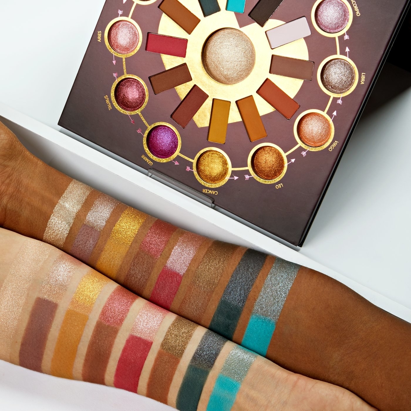 Two arms covered in eyeshadow swatches display the colors. The palette pans are arranged in a circle with the highlighter in the middle