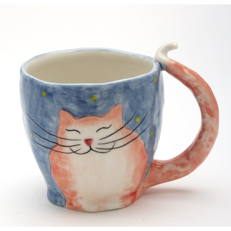 A porcelain mug hand-painted with a happy smiling reddish-orange cat on a blue background. The cat has large thin black whiskers and its tail acts as the mug&#x27;s handle, curving out at the top like a cat&#x27;s tail does