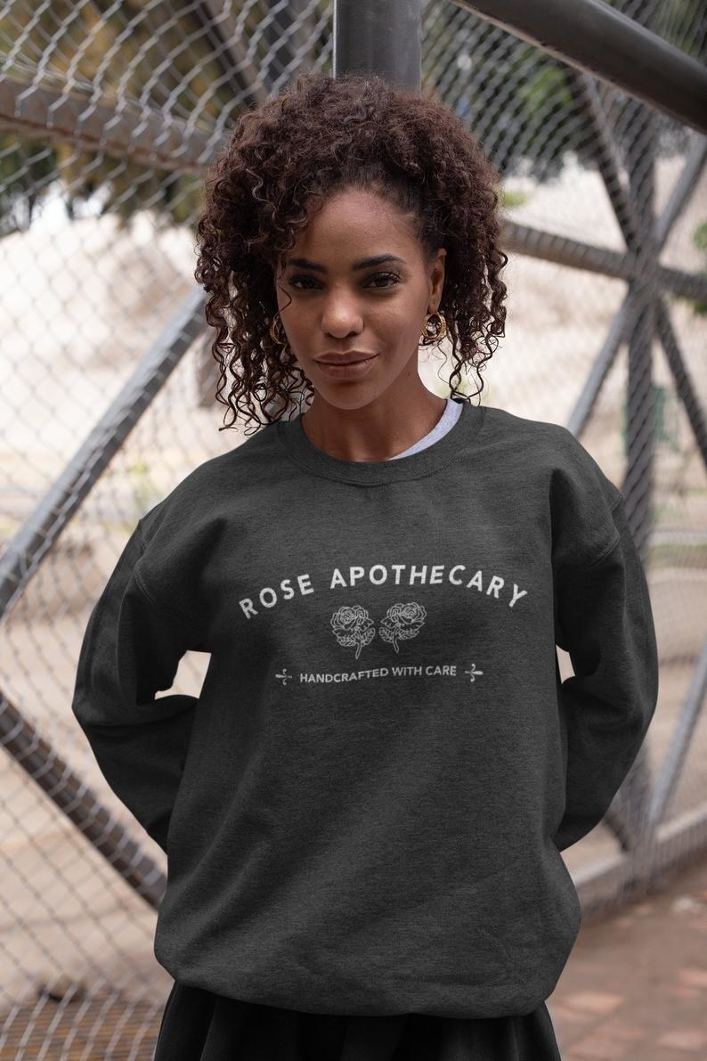 A person wearing a gray pullover sweater that that has a Rose Apothecary logo with two roses and the words &quot;handcrafted with care&quot; in small print under them 