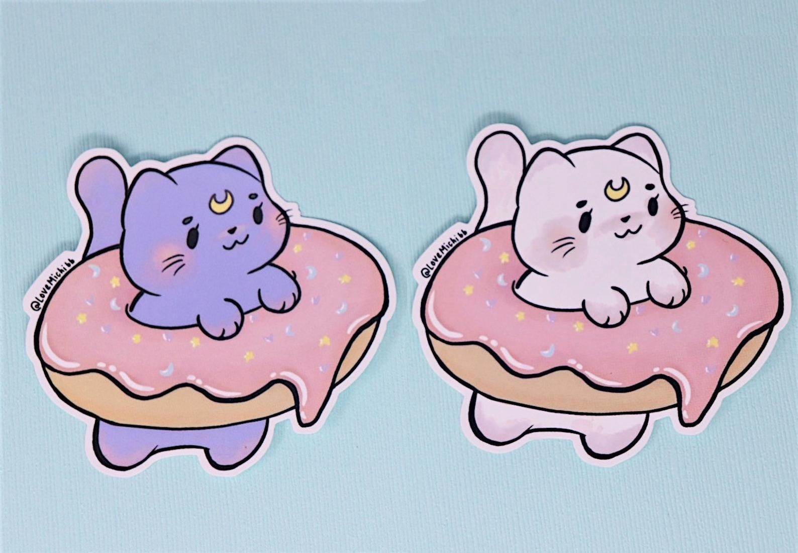 Two of the stickers, one pink cat and one purple cat, sitting in two donuts with pink frosting with sprinkles against a blue background