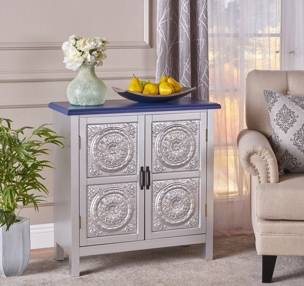Silver farmhouse-style cabinet with two front doors, blue tabletop, black drawer pulls, and a design on the cabinet doors that look like a tin-tile ceiling