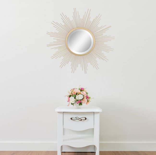 Sun-shaped wall mirror hanging above a white nightstand with a bouquet of flowers on top