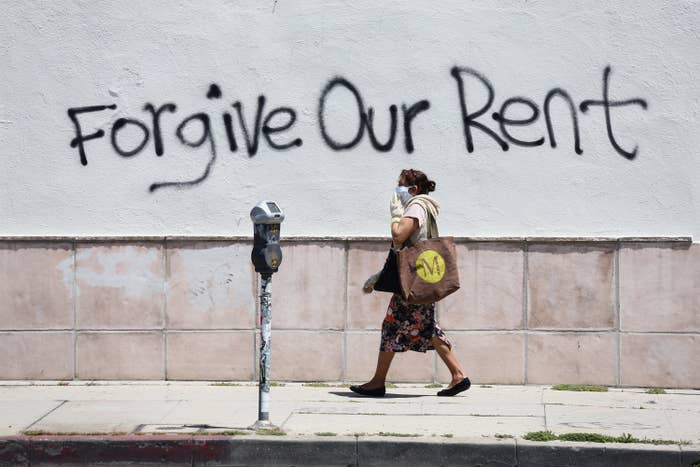 A woman wearing a mask walks past a wall with spray paint reading, &quot;Forgive Our Rent.&quot;