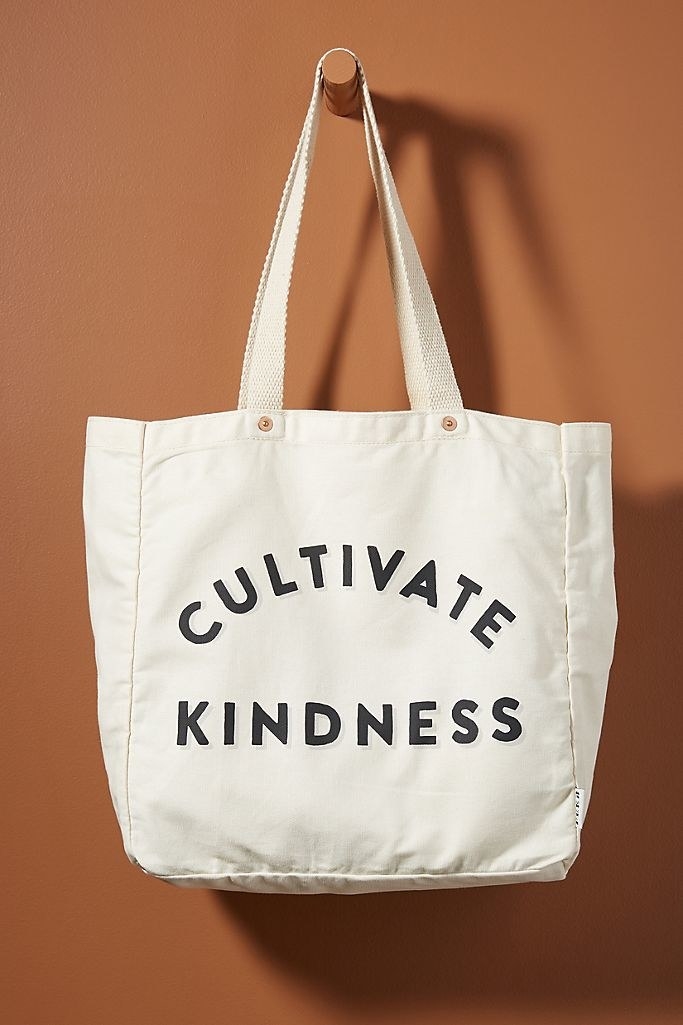 A cotton tote bag that says "Cultivate Kindness" hanging from a wooden hook on a brown wall