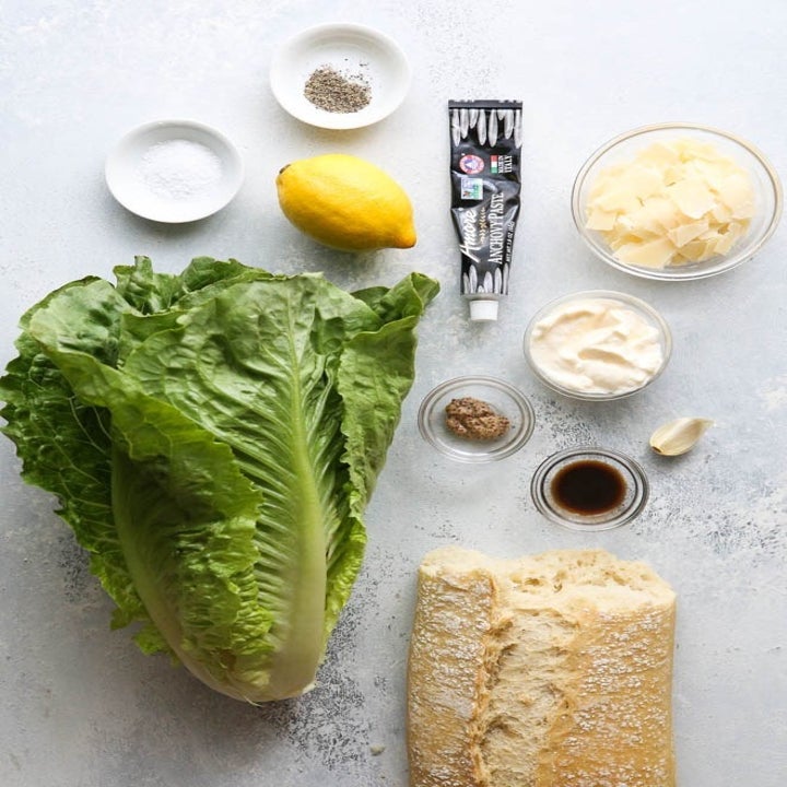 Ingredients for Caesar salad on counter including lettuce, lemon, anchovy paste, and grated Parmesan.