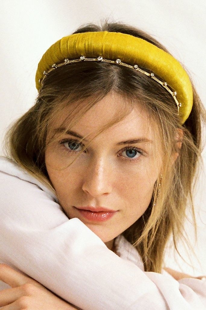 A model wearing a large gold satin headband, accessorizing with a smaller diamond headband and hair down