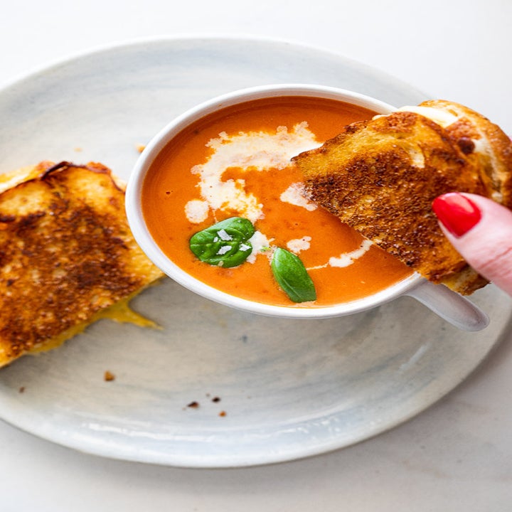 A hand dunking half of a grilled cheese sandwich into a cup of tomato soup 