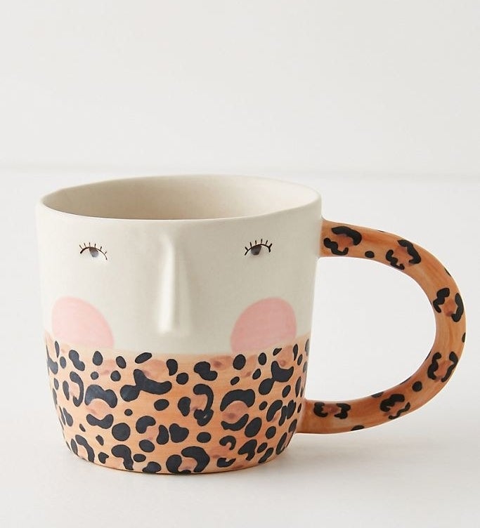 A wide handle mug with painted eyes, eyelashes, a sculpted nose, and painted cheeks. The bottom half is leopard print that is also seen on the handle