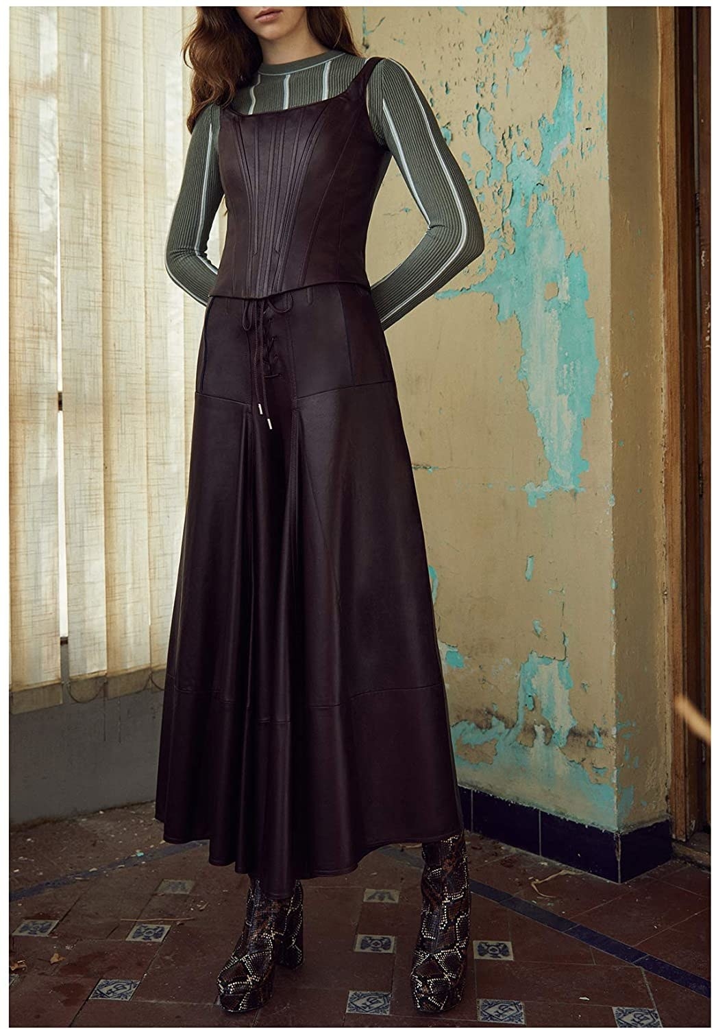 A model standing in a run-down room wearing the Alejandra Alonso Rojas INES Leather Culotte Pants with a matching top and platform snake print boots. 