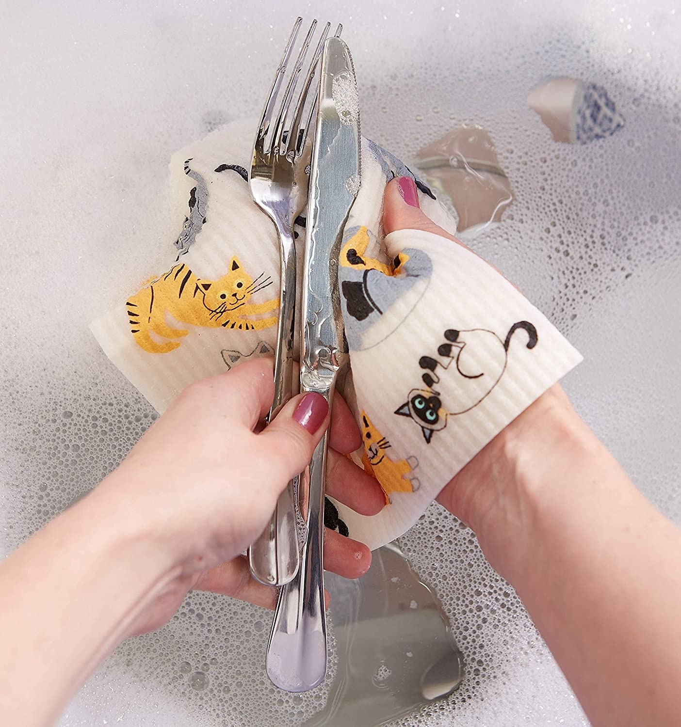 A person washing a knife and fork with a square-shaped Swedish dish cloth with happy cats printed on it. 