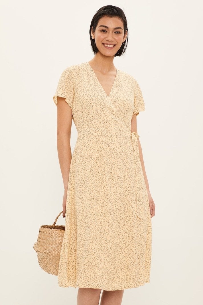 model in a yellow wrap dress with small darker yellow flower print. it ties at the side and has a v-neck and cap sleeves