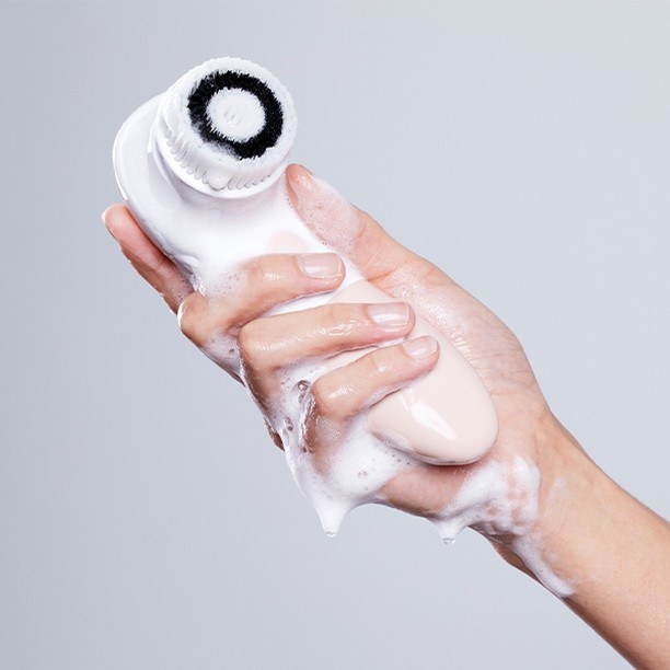 A model&#x27;s hand holding the facial cleansing brush that&#x27;s covered in foam