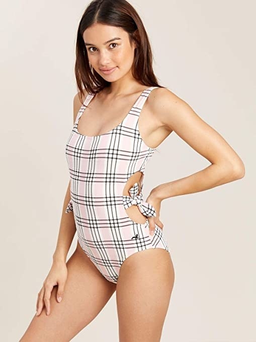 model against a beige backdrop wearing a one piece swimsuit in beige and pink plaid pattern. Suit has cutouts on the side with a tie in the midddle