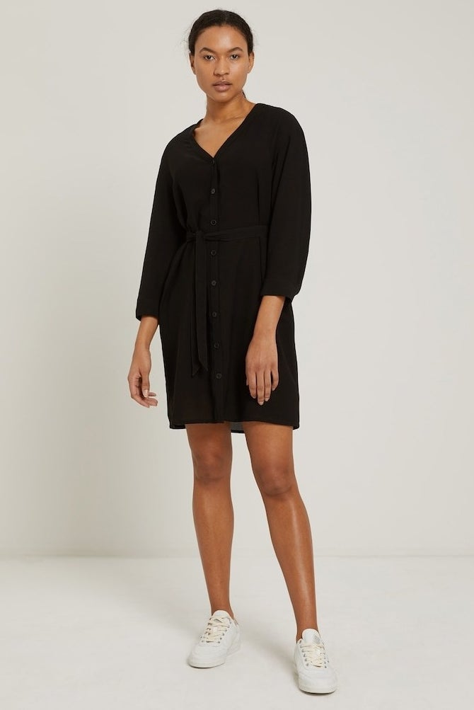 model in black v-neck dress with center buttons, a waist tie, and long sleeves that comes to the mid thigh, styled with white sneakers