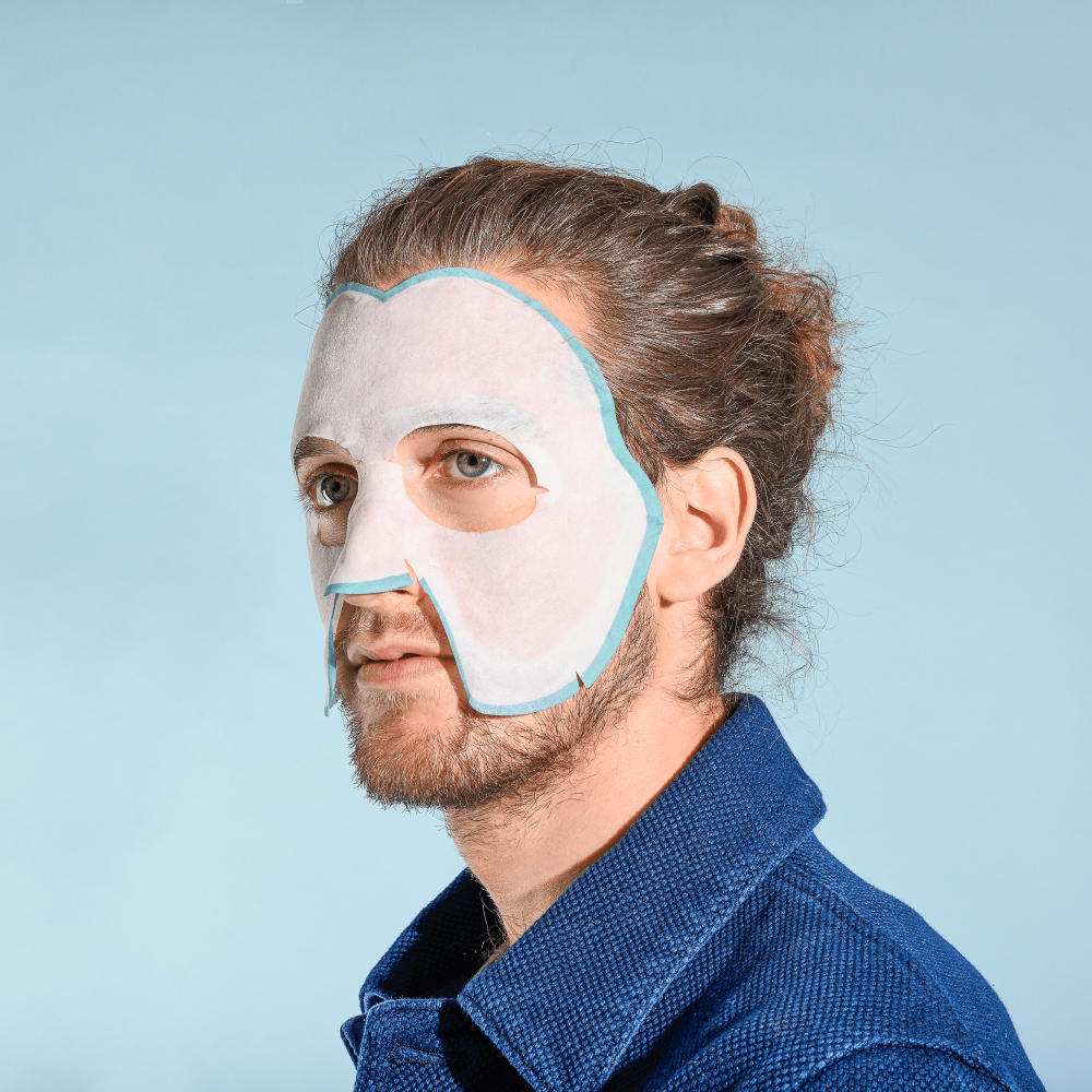 A model with a beard wearing the sheet mask; it covers all of the face except the eyes, upper lip, lips, lower jawline, and chin