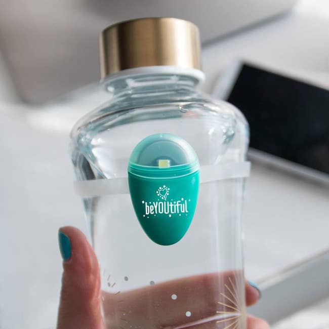 The tear-drop shaped hydration tracker hooked onto a silicone band, which is then attached to a glass water bottle a model is holding
