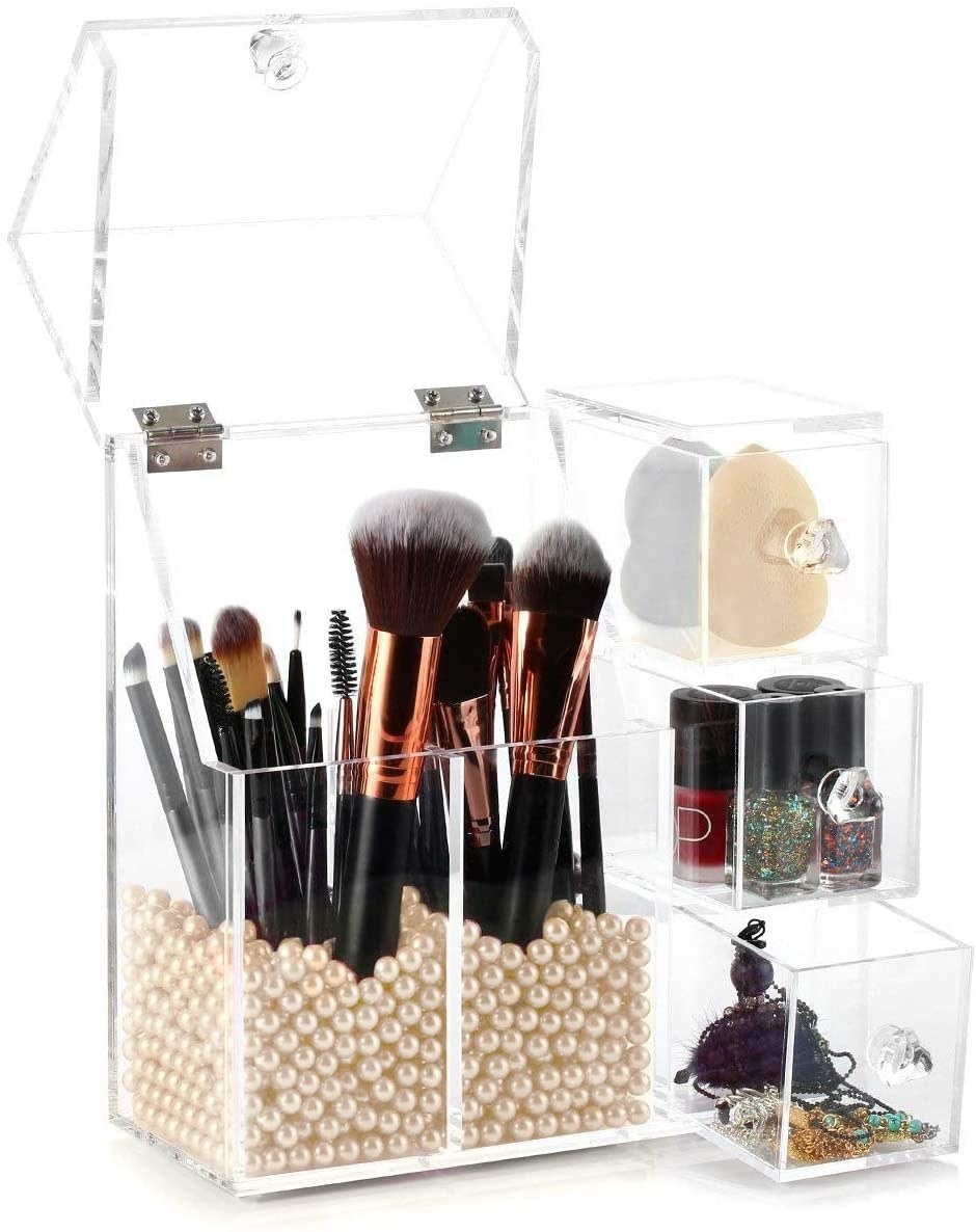 product shot of a clear organizer with two vertical storage compartments for brushes filled with beads at the bottom to keep them in place, three small drawers on the right side of the organizer
