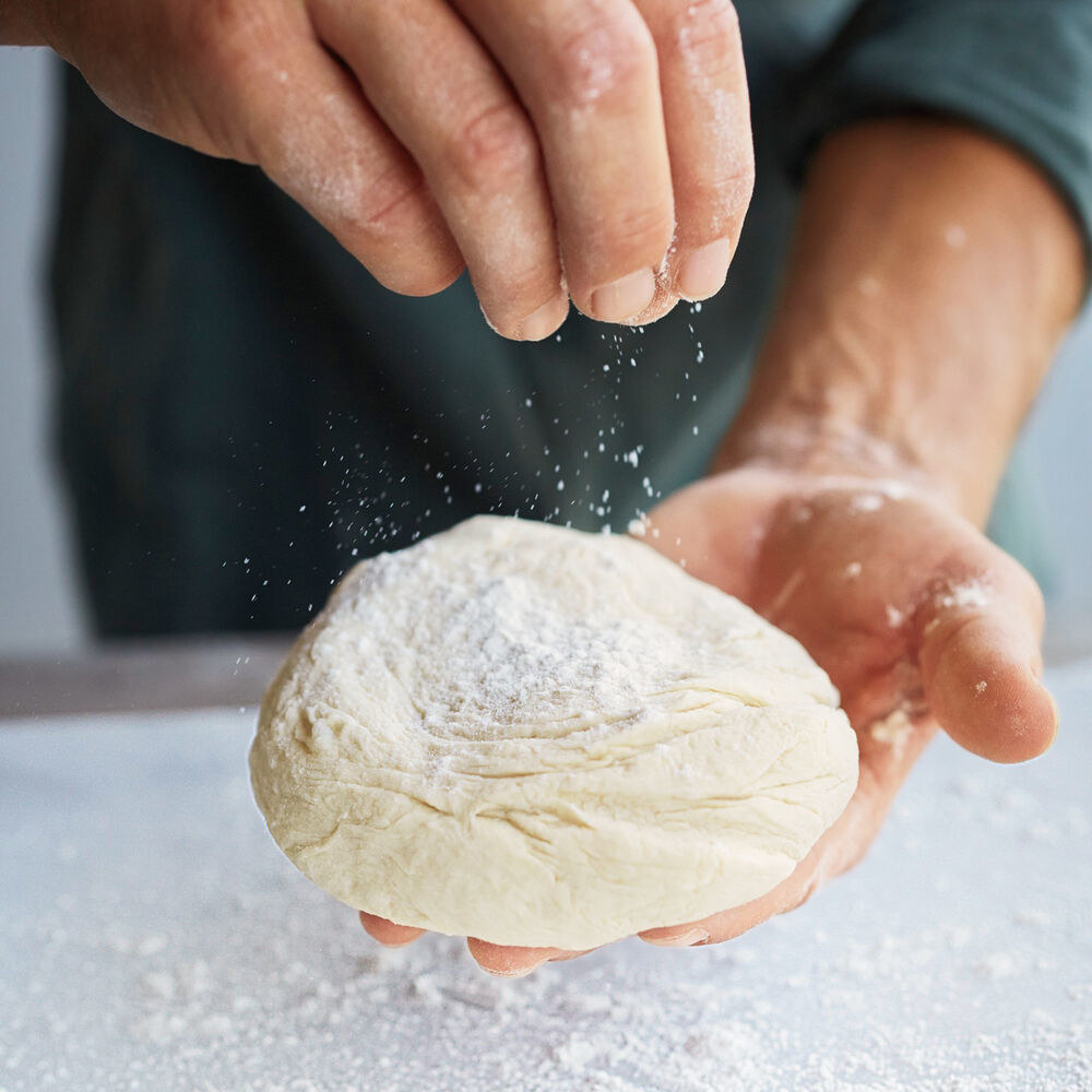 Hands sprinkling flour on a ball of pizza dough slightly larger than a tennis ball