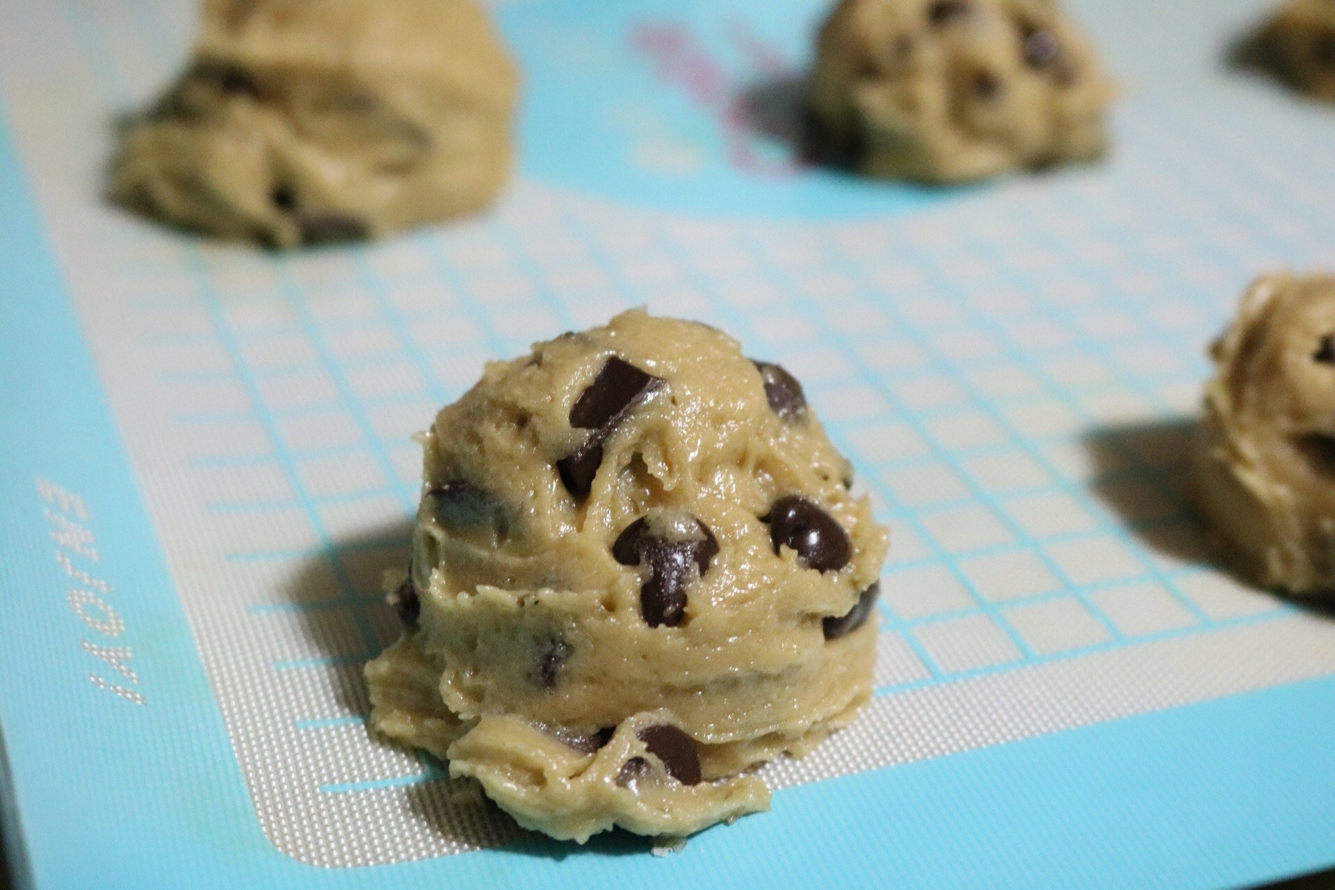A close-up of a round-shaped mound of cookie dough on a silicone baking sheet along with more mounds of cookie dough in the background