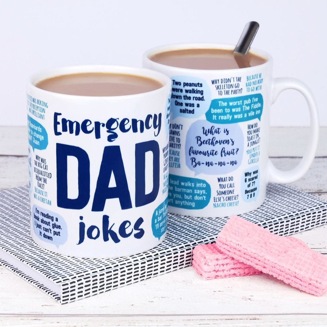 Two white mugs covered in blue text: The big letters say :emergency dad jokes&quot; and it is surrounded by word bubbles with punny jokes. The only visible joke says &quot;what is Beethoven&#x27;s favorite fruit? ba-na-na-na&quot;