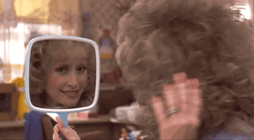 gif of Dolly Parton from the movie &quot;Steel Magnolias&quot; fixing her hair while looking into a hand mirror 