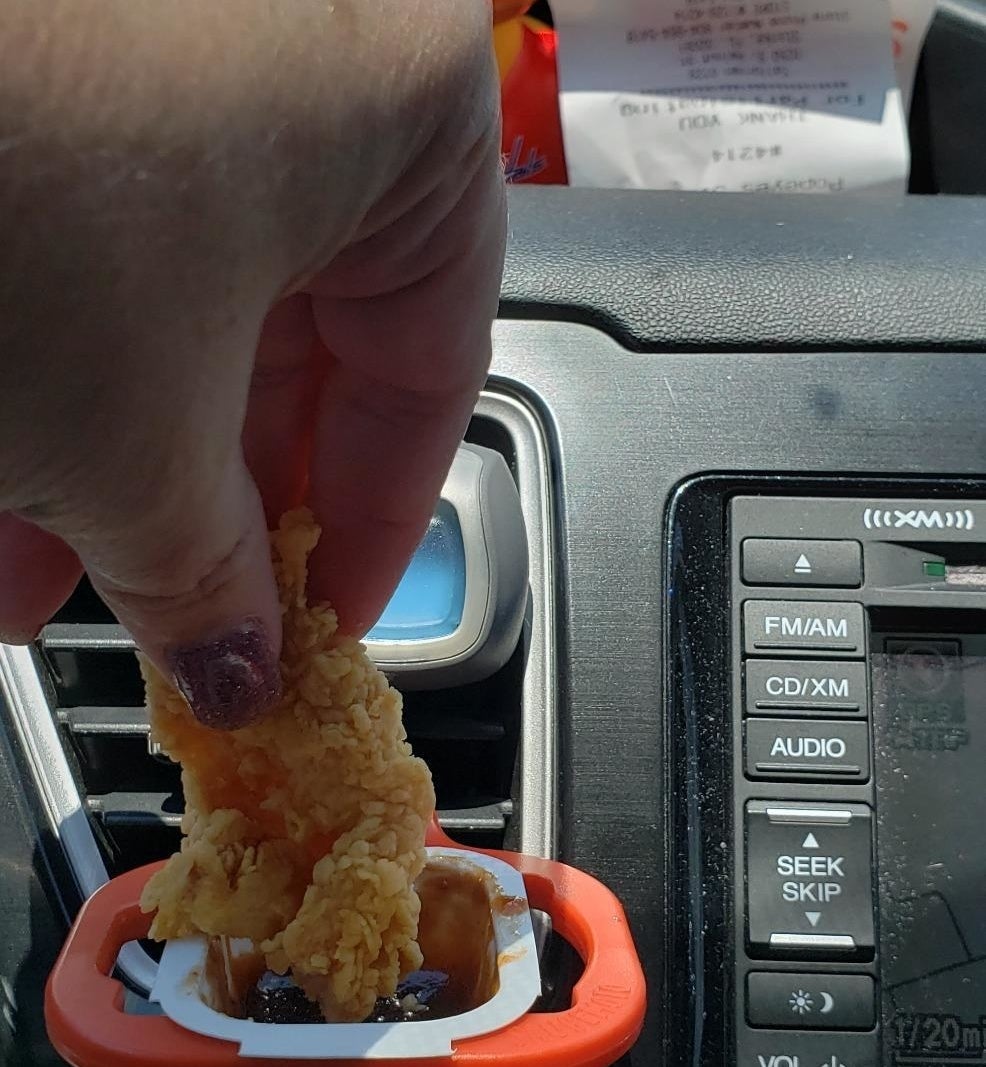 A person inside a car dipping a chicken tender into a sauce packet held in place by the dip clip, which is attached to the AC vent