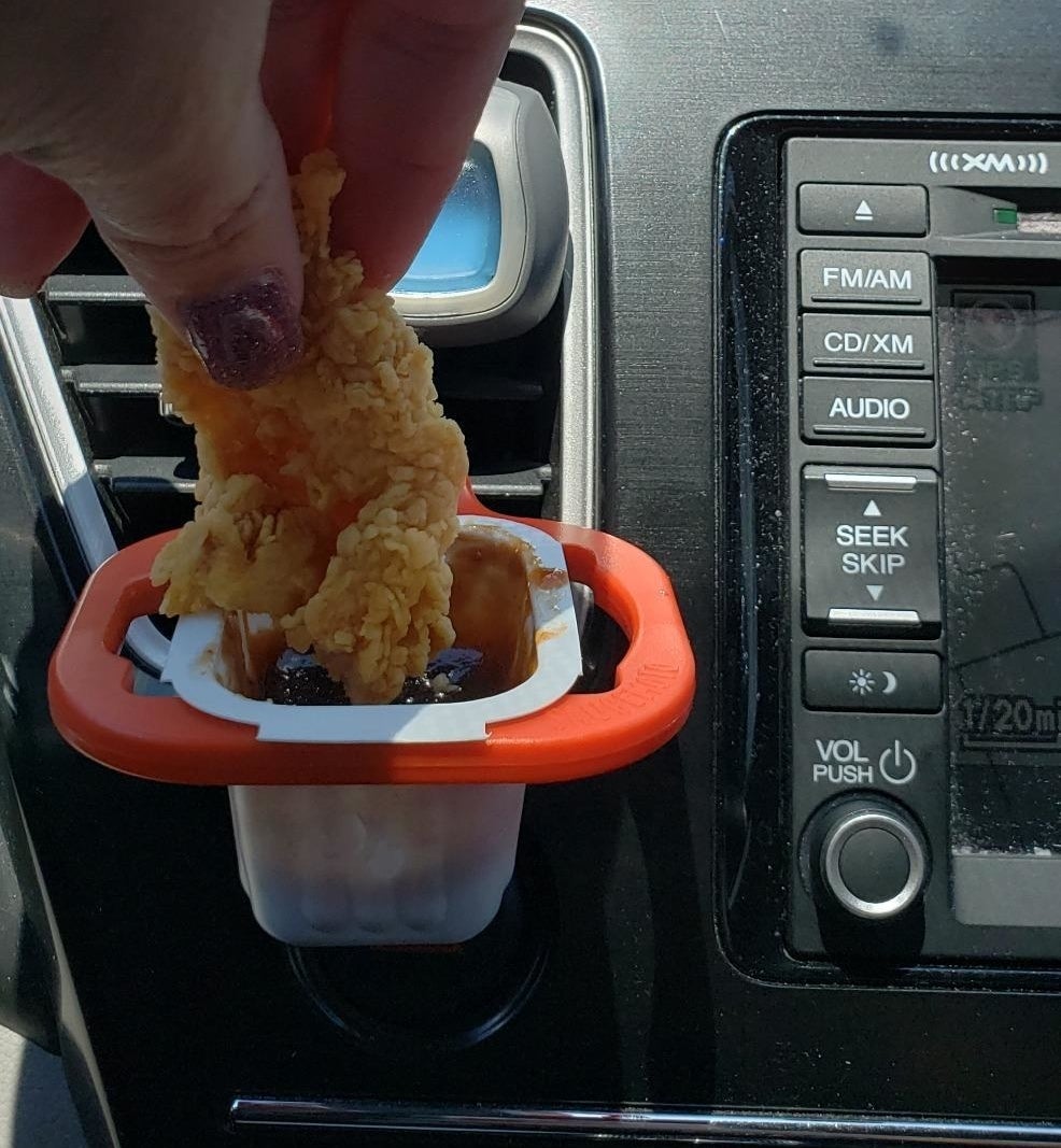 A person inside a car dipping a chicken tender into a sauce packet held in place by the dip clip, which is attached to the AC vent