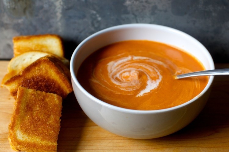 Grilled cheese beside a bowl of cream of tomato soup on a wooden board