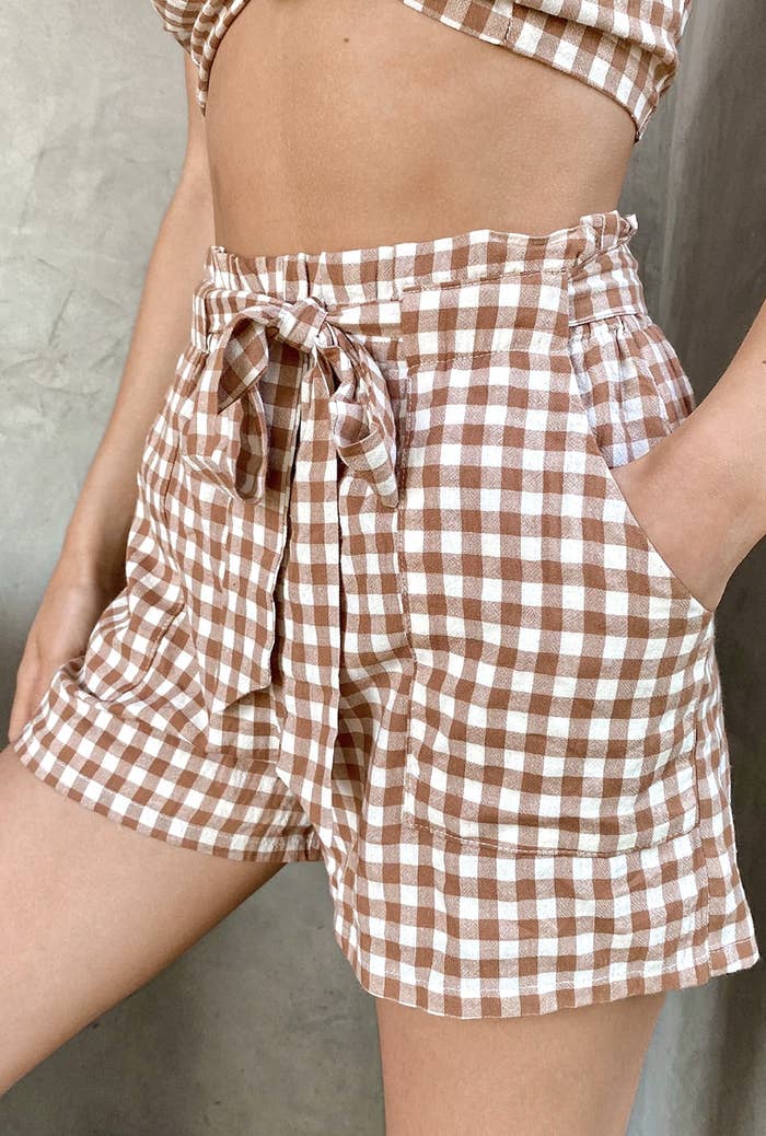 A close-up of a model wearing a pair of shorts with a brown and white checker pattern and a tie-waist front