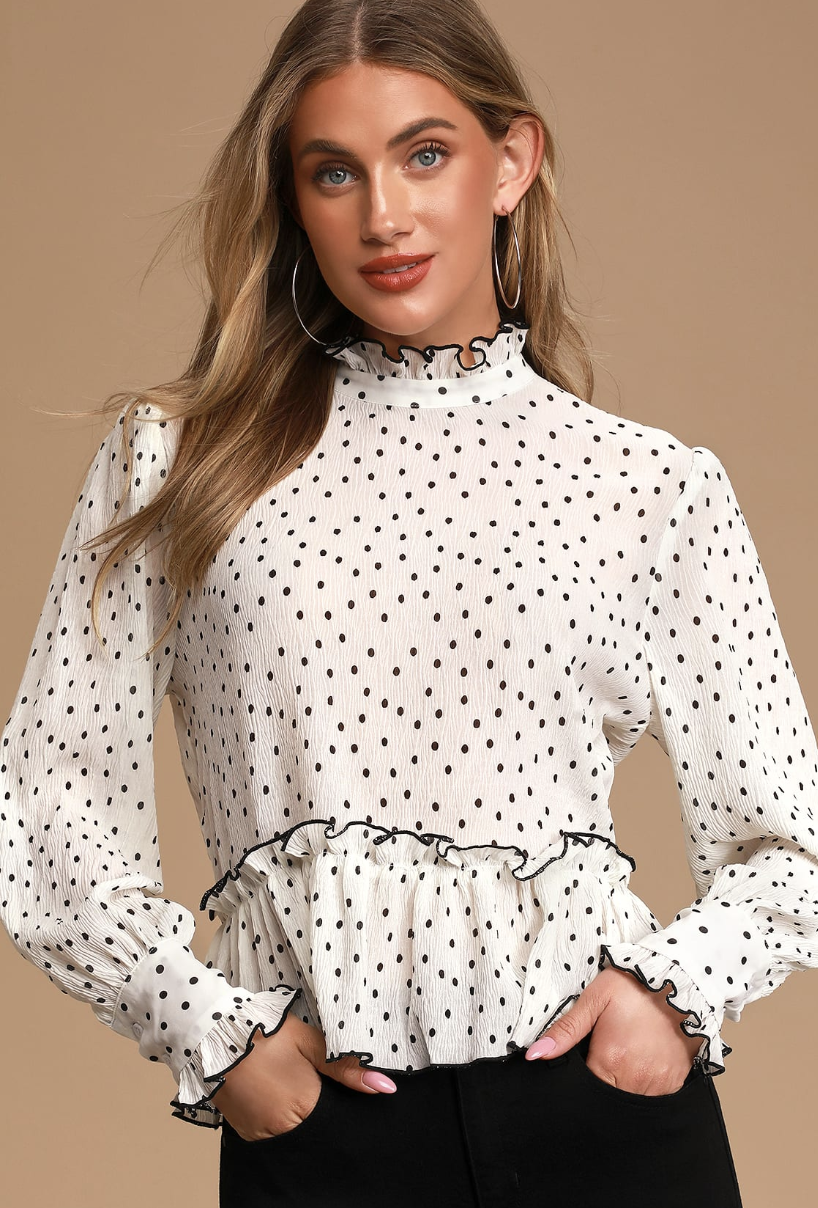 A medium shot of the model wearing the blouse, which is white with small black polka dots, has long sleeves, and has a ruffle detail at the waist and wrists