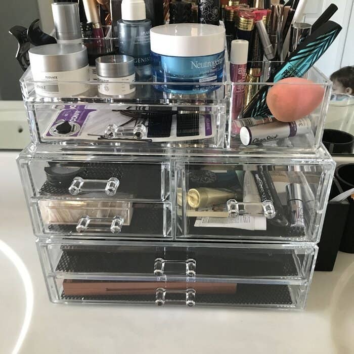A clear plastic cosmetic organizer with multiple drawers and compartments filled with various makeup products