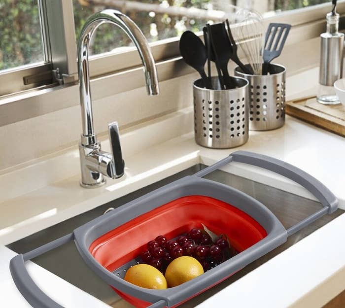A red plastic colander with gray arms extended and suspending itself with freshly washed fruit inside a sink