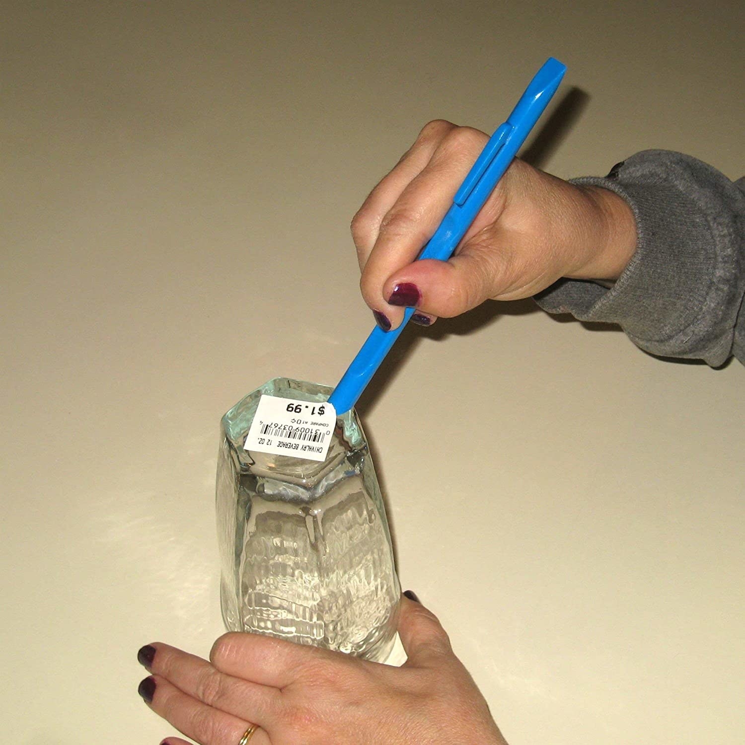 person with painted nails holding pen-like plastic tool in one hand, a glass in another hand, using the tool to scrape off a sticker on the bottom of the glass